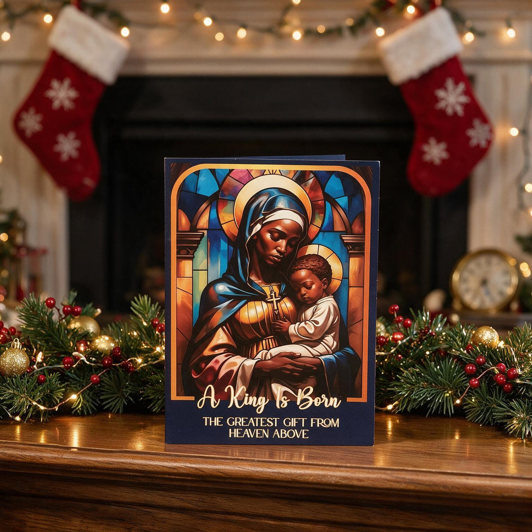 The Greatest Gift: African American Christmas Card Box Set by Janine Kai Robinson (Lifestyle 4) - A Christmas-themed greeting card standing on a wooden mantel, featuring a stained-glass-style illustration of Mary holding baby Jesus with the text 'A King Is Born' and 'THE GREATEST GIFT FROM HEAVEN ABOVE.' The background shows a cozy fireplace with stockings, greenery, and warm lights.