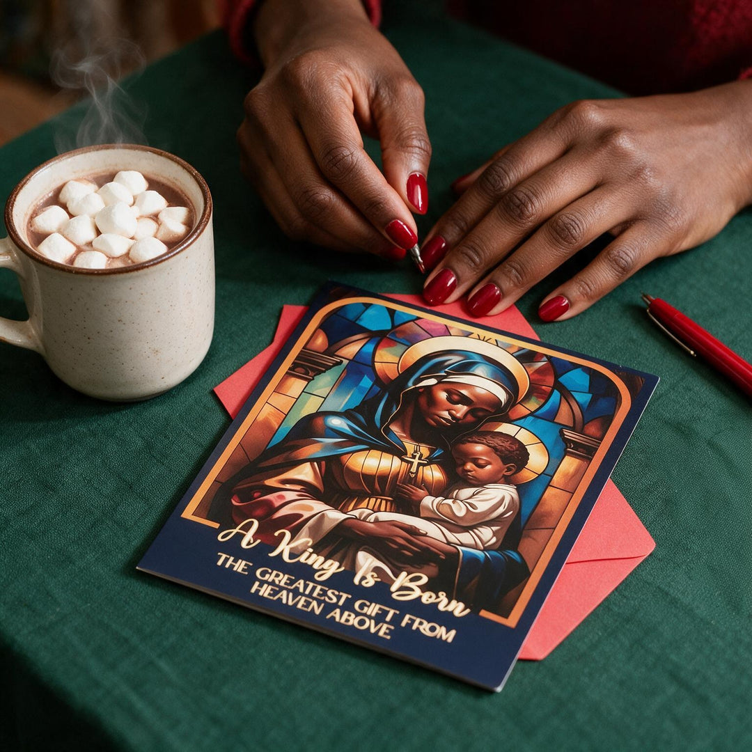 The Greatest Gift: African American Christmas Card Box Set by Janine Kai Robinson (Lifestyle 5) - A greeting card featuring a colorful religious illustration of Mary holding baby Jesus with the text 'A King Is Born' and 'THE GREATEST GIFT FROM HEAVEN ABOVE,' placed on a green table with a red envelope, a mug of hot chocolate with marshmallows, and a person's hands visible.