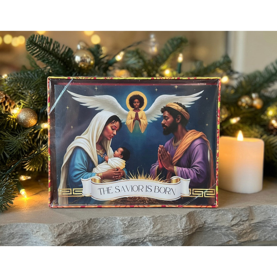 The Savior Is Born: African American  Christmas Card Box Set by Janine Kai Robinson (Lifestyle) - A boxed Christmas nativity print or card titled "THE SAVIOR IS BORN" sits on a stone surface with a lit candle and evergreen branches in the background. The front shows an illustrated scene of Mary, Joseph, and baby Jesus with an angel.