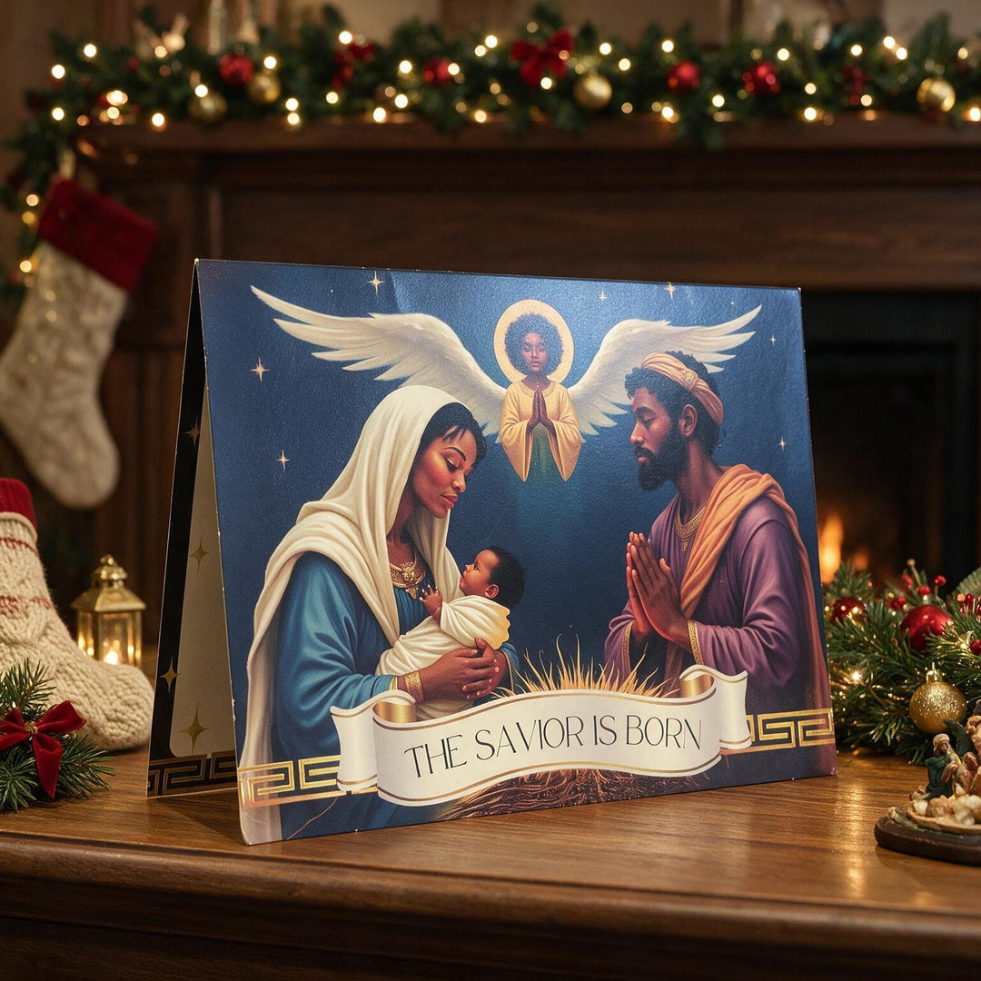 The Savior Is Born: African American  Christmas Card Box Set by Janine Kai Robinson (Lifestyle 2) - A Christmas nativity art print or card displayed on a wooden mantel, showing Mary, Joseph, baby Jesus, and an angel. The scene is decorated with a lit Christmas mantel, stockings, and garlands in the background.