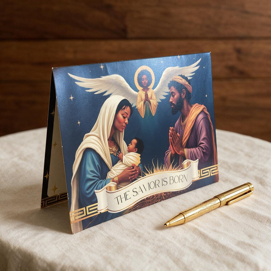 The Savior Is Born: African American  Christmas Card Box Set by Janine Kai Robinson (Lifestyle 3) - A folded greeting card standing on a light fabric surface, showing a religious illustration of Mary, Joseph, and baby Jesus under a white banner that reads 'THE SAVIOR IS BORN.' A gold pen rests beside the card.
