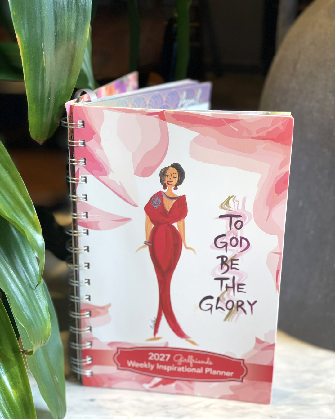 A spiral-bound 2027 Calliope Weekly Inspirational Planner standing upright. The cover features a white background with pink watercolor brushstrokes, an illustration of a woman in a red dress, and the text 'To God Be The Glory' and '2027 Calliope Weekly Inspirational Planner'.