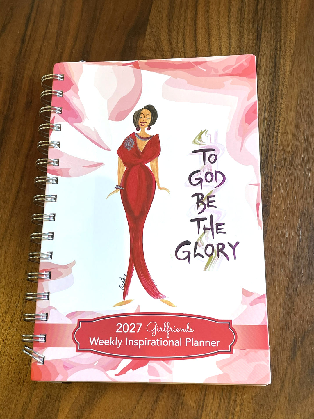 A spiral-bound 2027 Girlfriend Weekly Inspirational Planner lying on a wooden table. The cover features a white background with pink watercolor floral shapes, an illustration of a woman in a red dress, and the text 'To God Be The Glory' and '2027 Girlfriend Weekly Inspirational Planner'.