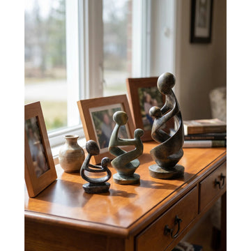 Ukama: Hand Carved Mother and Child Shona Sculpture (Lifestyle) - Four abstract family sculpture figurines in dark bronze and greenish-gray tones arranged on a polished wooden console table in a sunlit room, with framed photos and a small vase nearby.