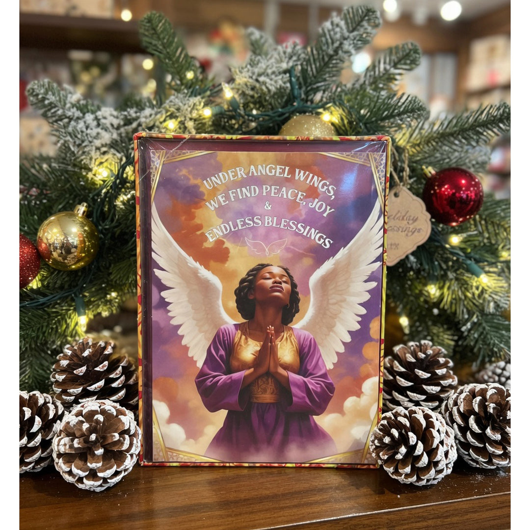 Peace, Joy and Endless Blessings: African American Christmas Card Box Set by Janine Kai Robinson (Lifestyle) - A boxed or book-style product standing upright on a wooden surface, surrounded by a decorated Christmas tree branch with pinecones and warm string lights. The front cover shows an angel with white wings in a purple robe, with the text 'Under Angel Wings We Find Peace Joy & Endless Blessings.